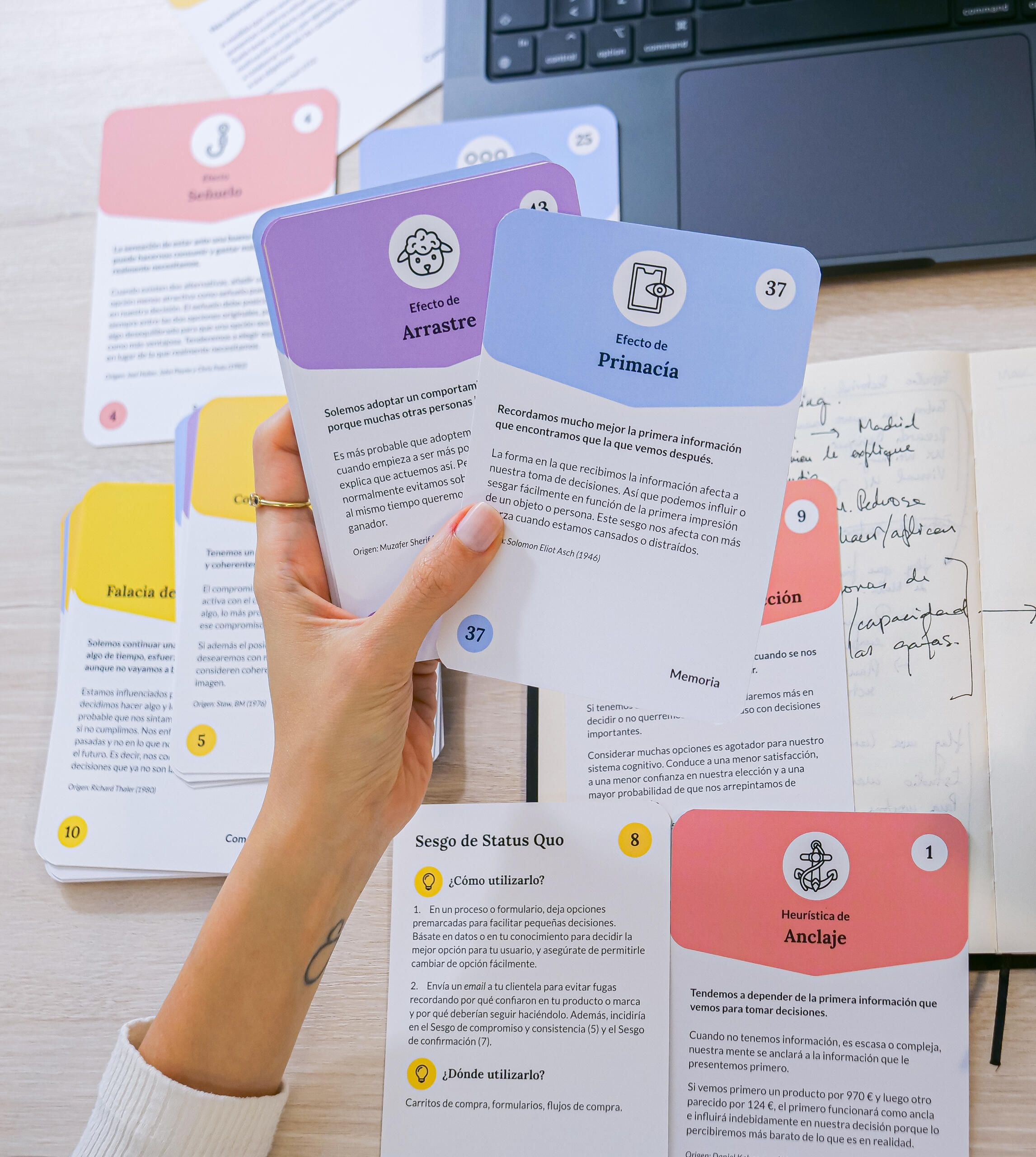 Imagen de las Behavioral cards Una mano en primer plano sostiene dos naipes: Efecto de arrastre (color lila) y Efecto de primacía (color azul) al fondo un portátil, apuntes en cuaderno y varios naipes dispersos
